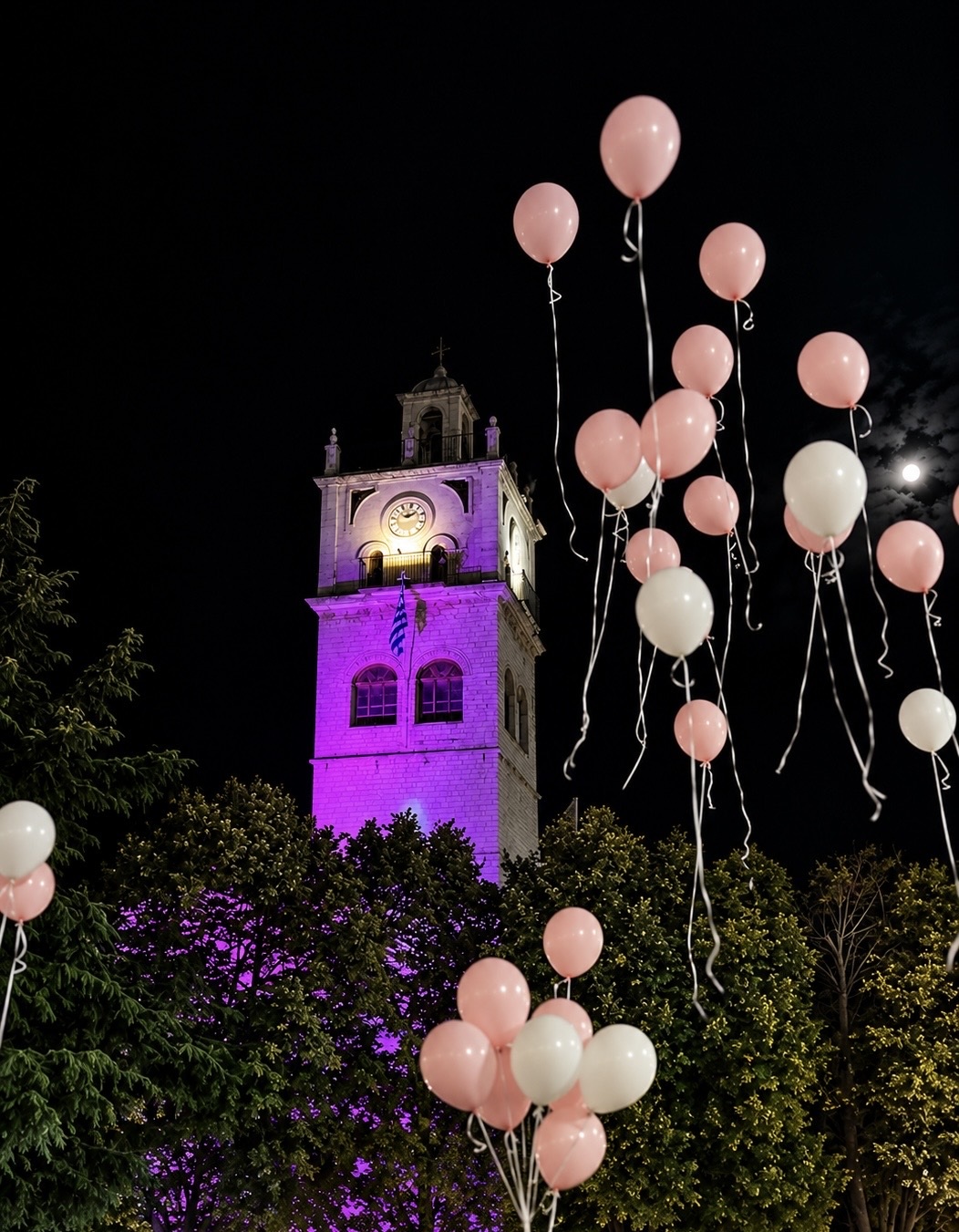 Ροζ και λευκά μπαλόνια δίπλα στο Ρολόι της Κοζάνης φωτισμένο σε μωβ χρώμα, στη βραδινή πορεία ευαισθητοποίησης για τον καρκίνο